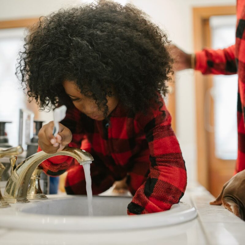 Father helping his young daughter brush her teeth at a bathroom sink, an example of ABA therapy daily living skills practiced at home